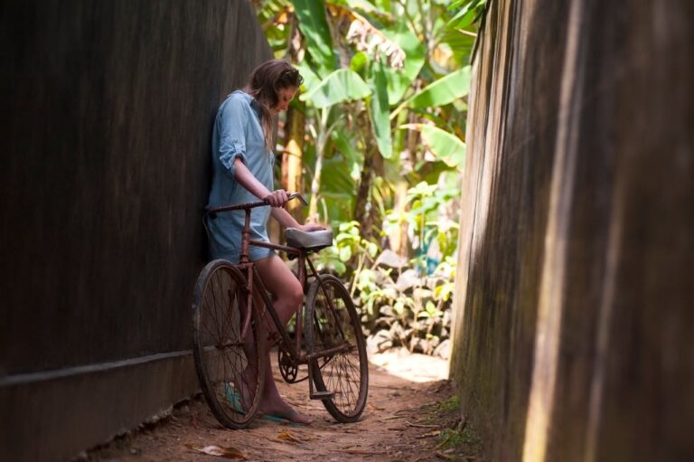 A young woman leans against a vintage bicycle in a tropical alleyway, enjoying a warm day outdoors.