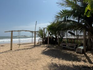 Sandy beach scene with swings, shaded bench, palm trees, and calm waves.