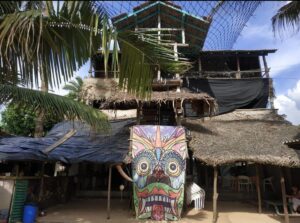 Multi-level tropical lodge with thatched roof, colorful tribal mask decor, palm trees, and shaded seating area.